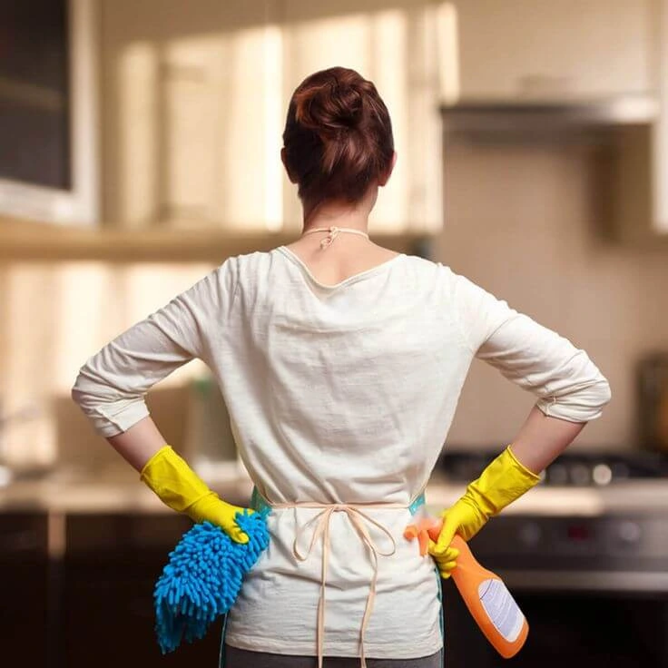 Back view of a professional cleaner wearing gloves and holding cleaning supplies in a modern kitchen.